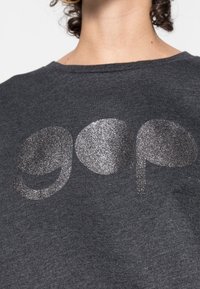 Persona vestida con una camiseta gris oscuro con la palabra "gap" impresa en grandes letras plateadas brillantes en el pecho.