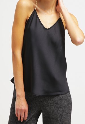 Kobieta ubrana w czarny satynowy bezrękawnik z cienkimi złotymi łańcuszkowymi ramiączkami oraz szare spodnie o fakturze, na jednolitym tle.