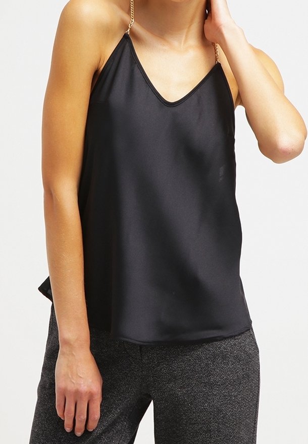 Femme portant un haut sans manches en satin noir avec de fines bretelles en chaîne dorée et un pantalon gris texturé sur un fond uni.