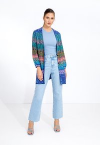 Knit cardigan with vibrant zigzag pattern in greens, blues, pinks, layered over light blue ribbed top. Paired with high-waisted jeans.
