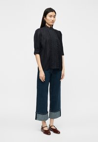 Γυναίκα που  στέκεται φορώντας μαύρο μπλουζάκι με δαντέλα, σκούρα τζιν με φαρδιά περιλαίμια και σκούρα καφέ παπούτσια Mary Jane, πάνω σε απλό λευκό φόντο.