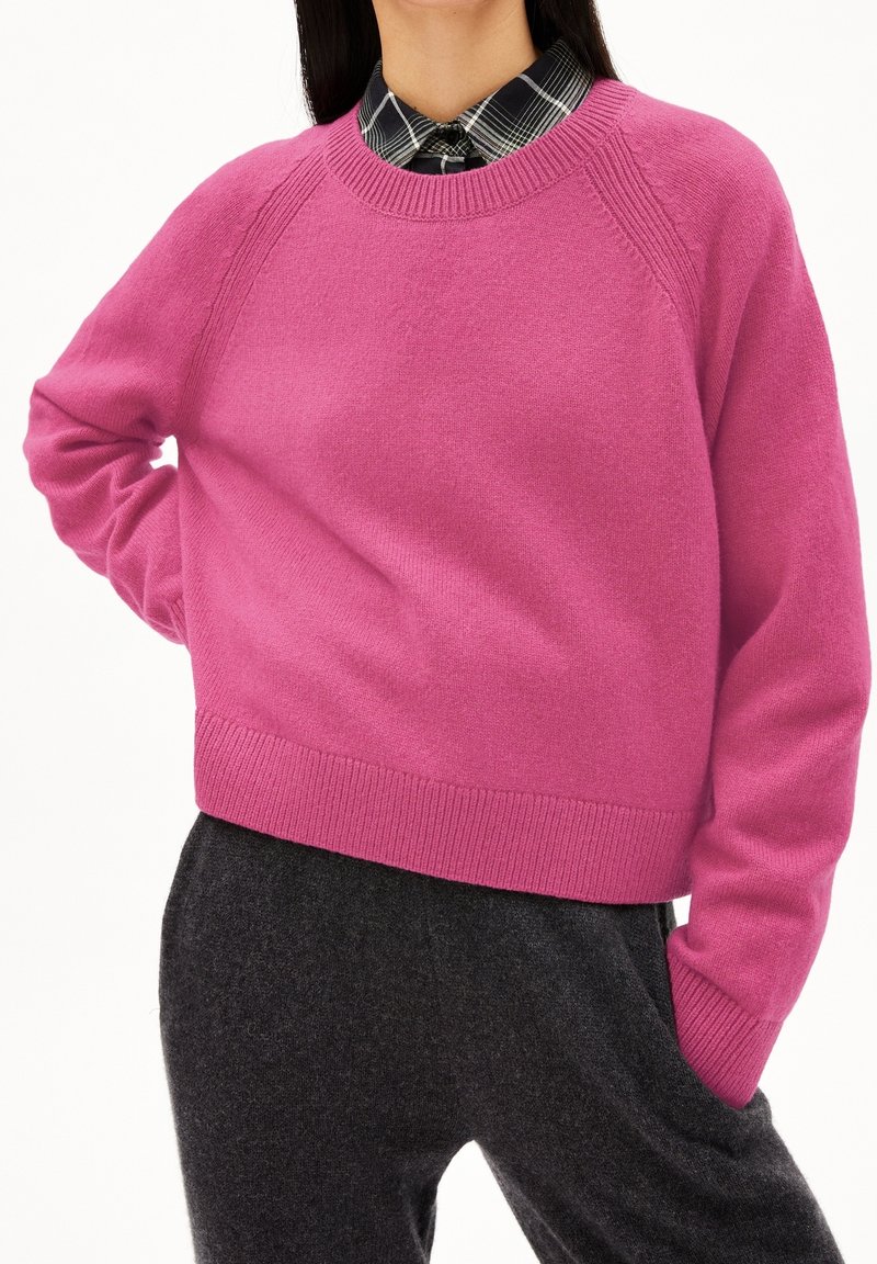 Pull rose avec un col rond côtelé et des manches raglan, associé à une chemise à carreaux noir et blanc. Le tissu semble doux et texturé.