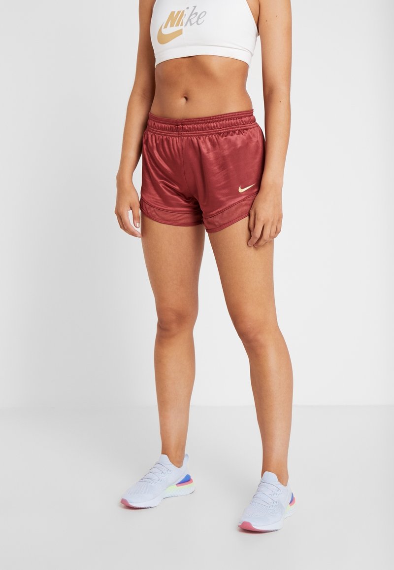 Femme portant un soutien-gorge de sport blanc Nike, un short Nike bordeaux brillant et des chaussures de course blanches avec des accents roses et bleus, debout sur un sol blanc.