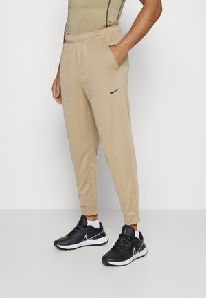 Man draagt beige Nike joggers met elastische tailleband en zwarte Nike sneakers, staande tegen een effen witte achtergrond.
