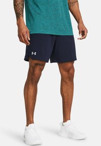 Mann in marineblauen Sportshorts mit Under Armour-Logo, einem türkisfarbenen gemusterten Hemd, weißen Turnschuhen und sichtbaren Tätowierungen an Beinen und Armen.