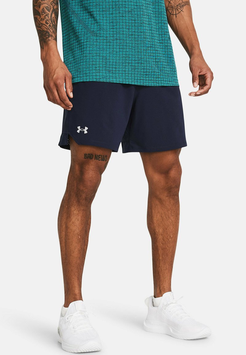 Mann in marineblauen Sportshorts mit Under Armour-Logo, einem türkisfarbenen gemusterten Hemd, weißen Turnschuhen und sichtbaren Tätowierungen an Beinen und Armen.