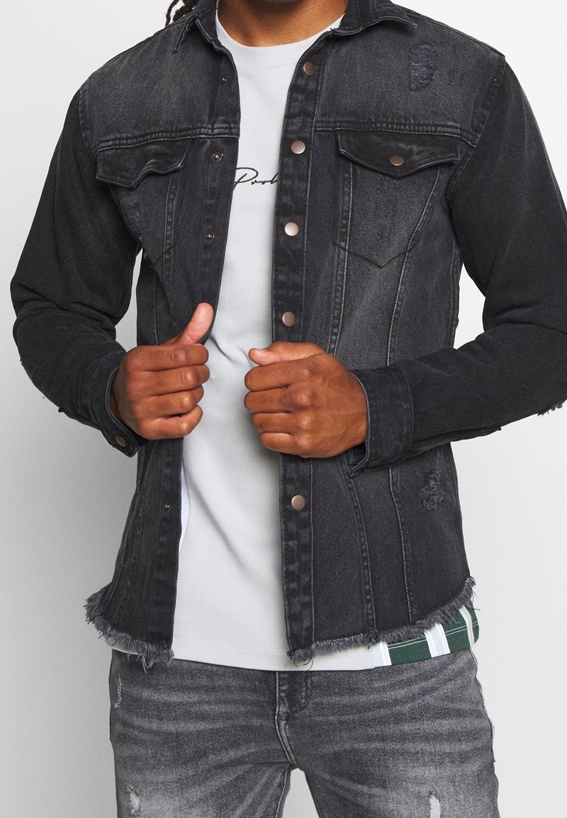 Personne portant une veste en denim noire avec des boutons métalliques sur un t-shirt blanc et un jean gris usé, tenant l'avant de la veste avec les deux mains.