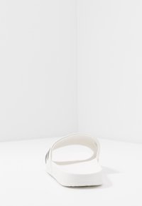 Sandalia plana blanca con una correa texturizada y una suela plana. Tiene un diseño minimalista con un acabado suave y bordes curvados.
