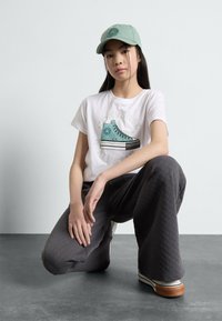 Camiseta blanca con un gráfico de unas zapatillas Converse verdes, pantalones acolchados grises y una gorra verde claro. El modelo está de rodillas sobre un fondo de suelo gris.