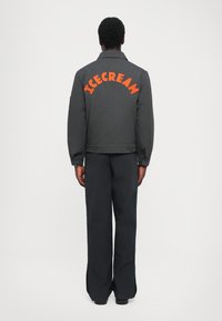 Zwarte cropped jas met oranje "ICECREAM" tekst op de rug, met knoopsluiting en brede mouwen; gecombineerd met bijpassende donkere broeken.