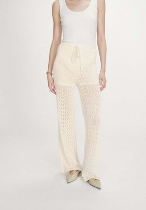 Femme portant un pantalon large en maille crème avec taille à cordon, un débardeur blanc côtelé, des chaussures beige à bout pointu et un bracelet en or.