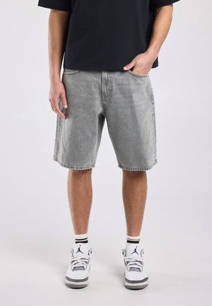 Man draagt grijze denim shorts, zwart T-shirt, witte hoge sneakers en witte sokken met zwarte strepen, staat met één hand in zijn zak.