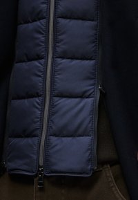 Μπλε σκούρο καπιτονέ μπουφάν αμάνικο με φερμουάρ στο μπροστινό μέρος, με οριζόντιες ραφές και λείο ύφασμα. Συνδυάζεται με σκούρο βασικό στρώμα και καφέ παντελόνι.