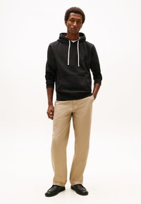 Fiatal férfi áll, kezét bézs nadrágja zsebében tartva, fekete kapucnis pulóvert visel fehér zsinórokkal és fekete cipőket egyszerű háttér előtt.