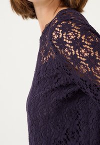 Haut en dentelle violet foncé avec des motifs floraux complexes, une texture transparente et un col rond avec des coutures détaillées le long de l'épaule.