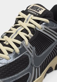 Pantof sport Nike Vomero 5 negru și gri, cu șireturi crem și partea superioară din plasă texturată, evidențiind cusăturile detaliate și designul talpii.
