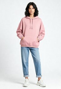 Felpa oversize color rosa chiaro con coulisse, tasche frontali, polsini e orlo a coste, abbinata a jeans azzurri rimbalzati e scarpe da ginnastica bianche.