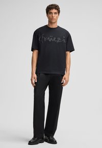 Schwarzes T-Shirt mit einem großen, tonalen Logo-Print auf der Vorderseite. Kurze Ärmel, lockere Passform. Trägt es mit schwarzen, locker sitzenden Hosen und schwarzen Schuhen.