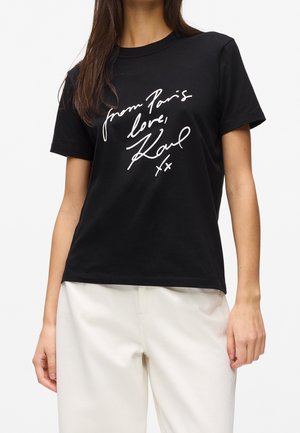 Kvinde iført en sort t-shirt med hvid tekst "from Paris love, Karl xx" kombineret med hvide bukser mod en ensfarvet baggrund.