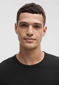 Mladý muž s krátkymi tmavými vlasmi, oblečený v čiernom tričku s okrúhlym výstrihom, pokojne sa pozerajúci do kamery na jednoduchom svetlom pozadí.