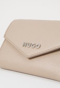 Portefeuille en simili cuir beige avec une surface texturée, présentant un logo "HUGO" en argent et une fermeture rabat.