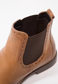 Bottine Chelsea en cuir marron avec panneau latéral élastique noir, détails brogue autour de l’ouverture et languette au talon.