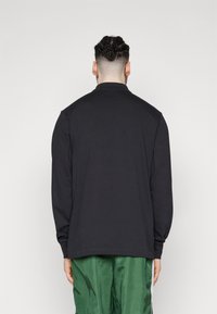 Camisa preta de mangas longas com gola redonda, corte solto e punhos canelados. Usada sobre calças verdes, exibindo uma textura suave e macia.