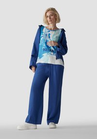 Leichter blauer Stoffanzug mit einer locker sitzenden Jacke und weiter Hose, kombiniert mit einem bedruckten T-Shirt, das eine blau-weiße Grafik zeigt.