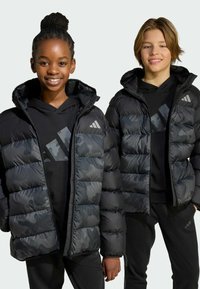 Deux personnes portant des vestes matelassées en camouflage noir avec capuche, présentant des sections matelassées contrastées et un branding subtil d'Adidas sur la poitrine.