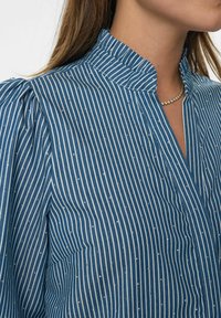 Zila svītrainā blūze ar stāvošu apkakli, kurai ir metāliski akcenti un vertikālas līnijas. Ir redzama maiga tekstūra un pūkainas piedurknes.