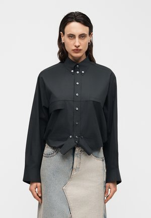 Persona con cabello oscuro que lleva una camisa negra abotonada metida en una falda vaquera bicolor, de pie contra un fondo claro y liso.