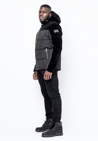 Puffjacka med svart slät tyg och plyschsvarta ärmar, fickor med dragkedja och huva, tillsammans med svarta slim fit-jeans och stövlar.