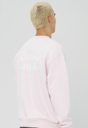 Sweat-shirt rose clair avec col rond, manches longues et un design en relief de texte blanc au dos indiquant "Édition Limitée hors série."