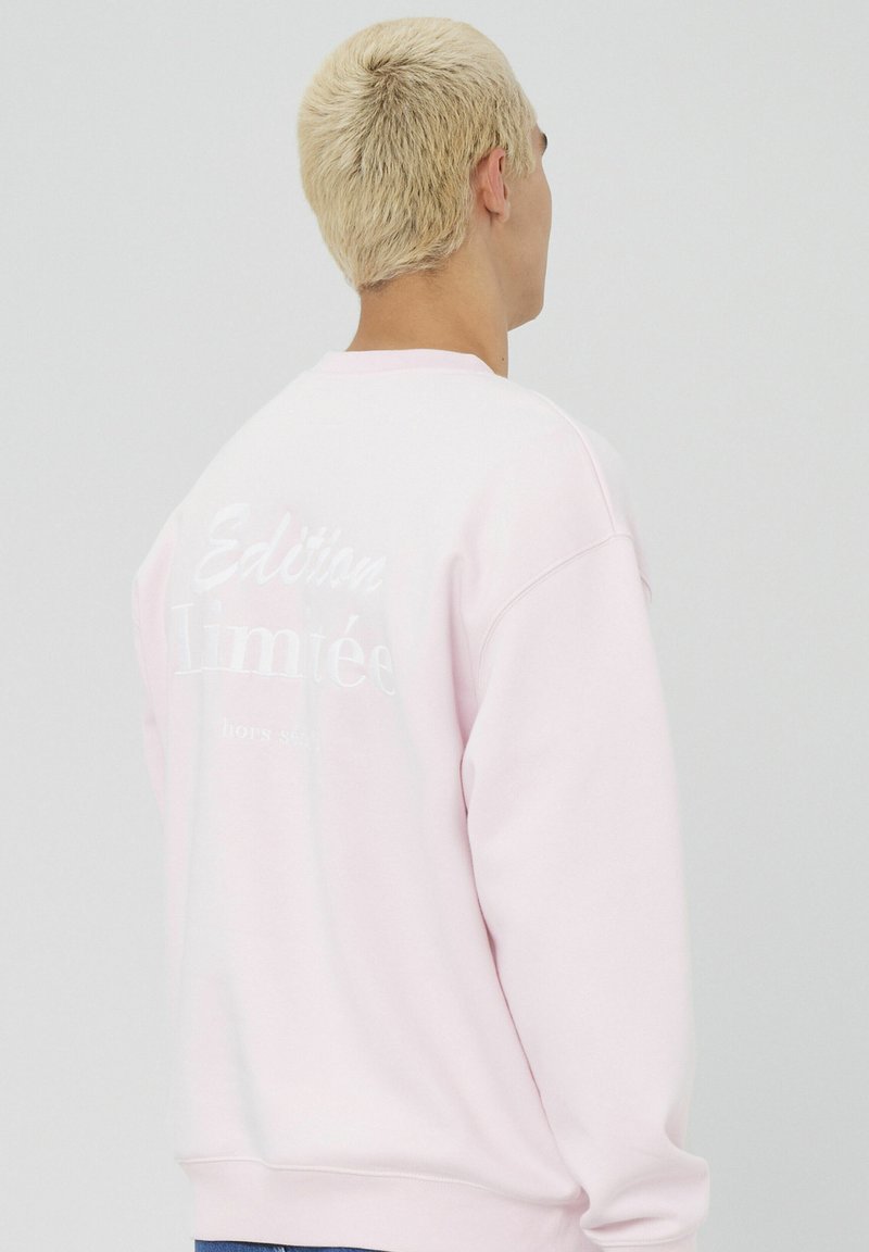Sweat-shirt rose clair avec col rond, manches longues et un design en relief de texte blanc au dos indiquant "Édition Limitée hors série."