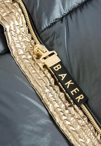 Chaqueta de plumas negra con cremallera y ribetes dorados brillantes. El tirador de la cremallera tiene grabado el nombre de la marca "Baker" en una etiqueta de tela negra.