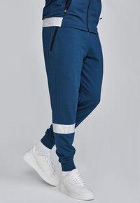 Marineblaue Jogginghosen aus weichem Material, mit einem weißen, horizontalen Streifen und Text an der Wade, kombiniert mit weißen Turnschuhen.