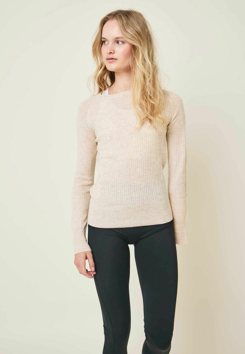 Jeune femme aux longs cheveux blonds portant un pull beige côtelé et des leggings noirs, regardant vers sa gauche sur un fond clair.