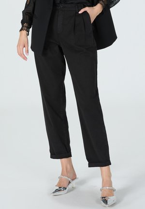 Hochtaillierte schwarze Baumwollhose mit Falten, schmal zulaufenden Beinen und umgekrempelten Säumen, kombiniert mit glänzenden silbernen spitz zulaufenden Absätzen.