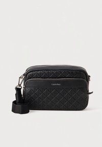 Black quilted leather Calvin Klein crossbody bag with front zipper pocket and adjustable fabric strap on white background.