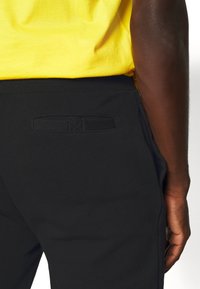 Sorte sweatpants med en glat tekstur, elastisk talje og baglomme med et vævet labeldesign. Lys gul skjorte delvist synlig.