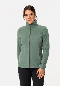 Giacca in pile verde con alto colletto, zip completa e due tasche laterali. Presenta una texture liscia e un logo del marchio discreto sul petto.