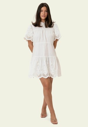 Moteris stovi sukryžiavusi kojas, dėvėdama baltą trumpomis rankovėmis suknelę su akių raštais ir kreminės spalvos basutes ant šviesaus fono.