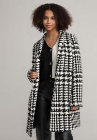 Schwarz-weißer Hahnentrittmantel mit lockerer Passform, ausgestattet mit einem breiten Revers und Knöpfen. Getragen über einem schwarzen Strickpullover und schwarzen Hosen.