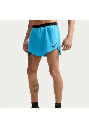 Mężczyzna w jaskrawo niebieskich spodenkach do biegania Nike z czarnym pasem i czarnymi skarpetkami, z widocznymi tatuażami na lewej nodze.