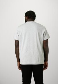 Tricou gri din bumbac, cu mâneci scurte, guler rotund și o croială lejeră, văzut din spate. Pantaloni negri sunt parțial vizibili.