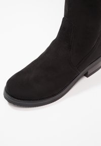 Bottine noire en daim avec bout rond, détail de couture, panneau latéral élastique et talon bas en caoutchouc sur fond blanc.