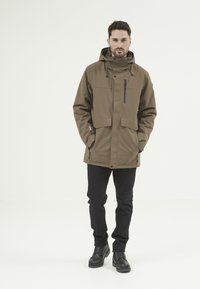 Brun vattentät jacka med stora framfickor, justerbar huva, dragkedja och tryckknappar, tillsammans med svarta byxor och stövlar.