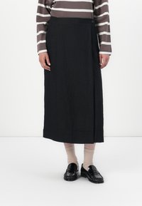 Svart, ankel-lang skjørt med innpakket design og knappdetalj i livet; kombinert med svarte loafers og sokker i nøytrale farger.