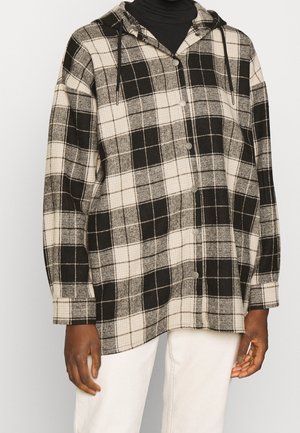 Zwart-wit geruit overhemd met knopen en capuchon, gedragen over een zwarte coltrui, gecombineerd met gebroken-witte broek, getoond op een staande persoon.