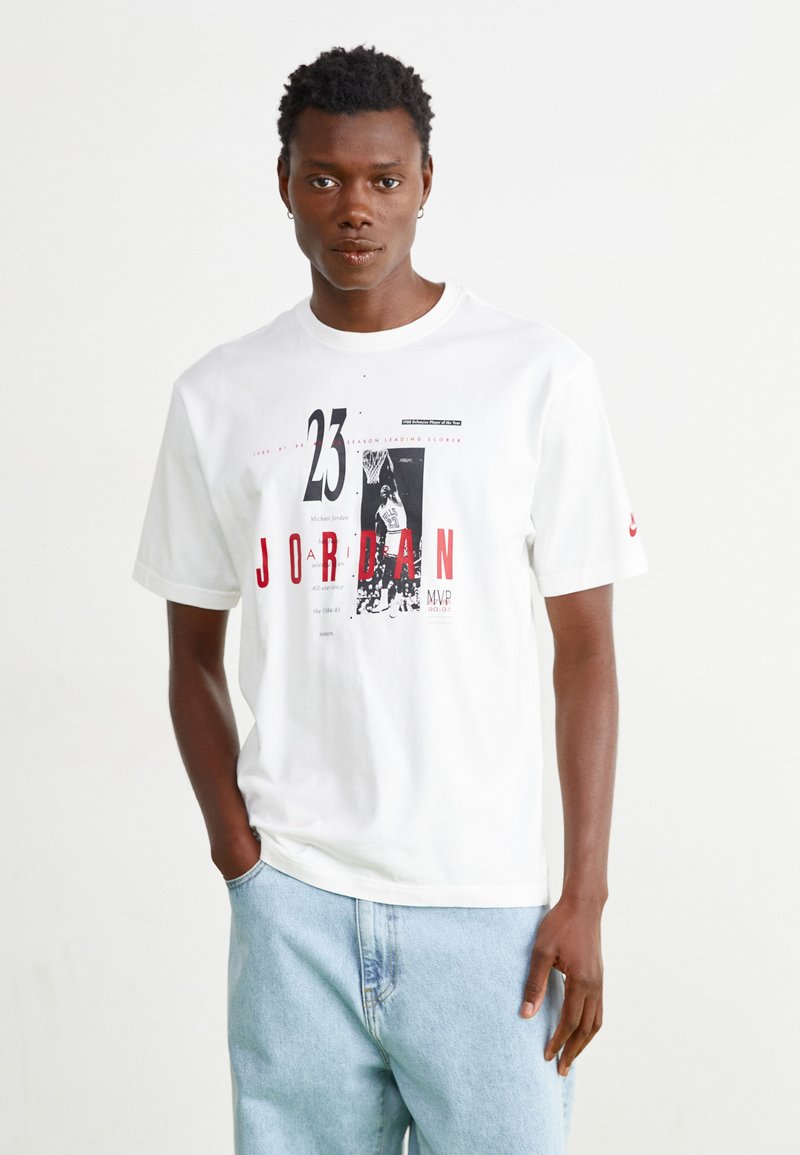 Vit bomullströja med grafisk tryck som har "JORDAN" och spelar nummer 23. Kort ärm, rund hals, avslappnad passform, i kombination med ljusblå jeans.